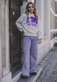 Grijze sweatshirt met paarse tekst "ST. TROPEZ FEEL THE VIBES," gecombineerd met lichtpaarse wijde jeans en zwarte schoenen. Buitensetting.