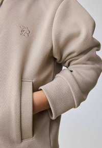 Beiger Kapuzenpullover mit strukturierter Stoffoberfläche, versehen mit einem kleinen gestickten Logo auf der Brust, einer Kängurutasche und umgeschlagenen Bündchen.