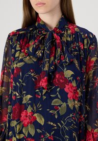 Floral blouse in sheer navy fabric with red and green floral patterns. Features a bow tie collar and pleated detailing.