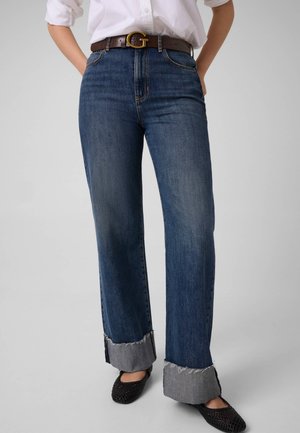 Un mannequin porte un jean bleu foncé à jambes larges avec de grands revers, une chemise blanche, une ceinture foncée à boucle dorée et des chaussures plates noires.