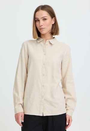 Mujer con cabello lacio castaño claro, lleva una camisa beige de manga larga con botones y pantalones negros, de pie frente a un fondo claro y liso.