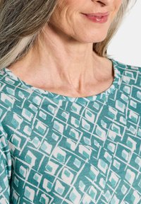 Türkise gemusterte Stoff mit weißen geometrischen Motiven. Das Design verfügt über eine weiche, glatte Textur und einen runden Halsausschnitt.