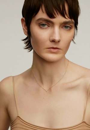 Collar de oro con una cadena delicada y un pequeño colgante triangular, utilizado sobre un hombro desnudo con una blusa sencilla de color claro.
