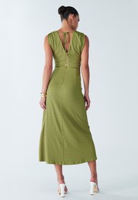 Olijfgroene midi-jurk met een getailleerd lijfje, achterritssluiting en een strikdetail bij de hals. Gladde stof met een subtiele textuur, gecombineerd met witte hakken.