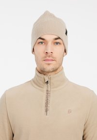 Beanie lavorato a maglia beige con punta, caratterizzato da una texture a costine. Indossato con un top zip-up in fleece beige abbinato con logo.