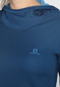 Blå träningshoodie med strukturerad tyg, figurnära design och justerbar huva. Har Salomon-logotypen på bröstet.