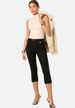 Schwarze, dunkel geschnittene Jeans mit Fronttaschen und goldenen Knopfakzenten. Das Model trägt ein beiges ärmelloses Oberteil und hält eine beäge Jacke.