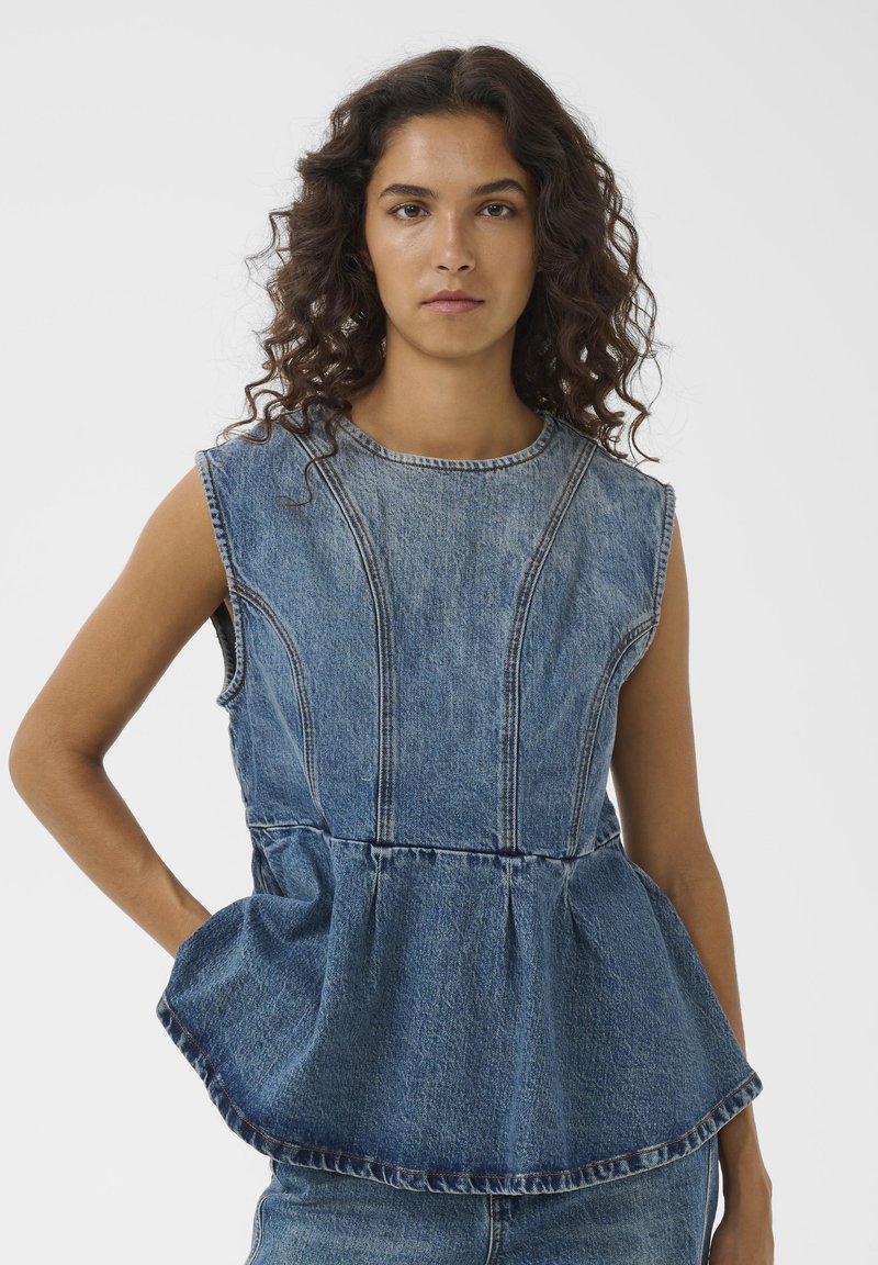 Femme aux cheveux bouclés portant un top péplum en denim bleu sans manches et un jean en denim assorti, debout devant un fond clair uni.