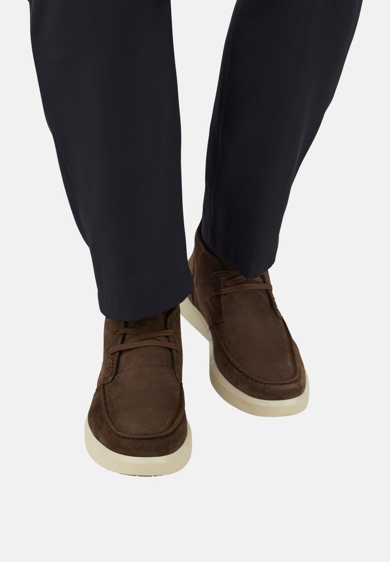 Zapatos informales de ante marrón con cordones, llevados con pantalones oscuros, mostrados desde una vista frontal en ángulo bajo sobre un fondo liso.