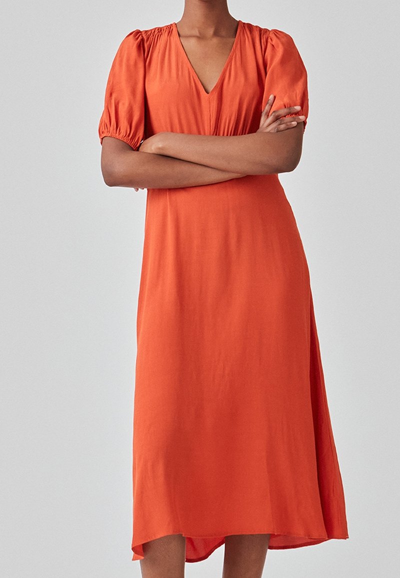 Robe orange mi-longue avec un col en V, des manches bouffantes et une coupe ample. Fabriquée dans un tissu doux à la texture lisse ; l'ourlet est légèrement plus long à l'arrière.