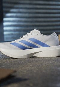 Scarpa sportiva bianca con tre strisce blu e suola spessa posizionata su una superficie strutturata con gradini sfocati sullo sfondo.