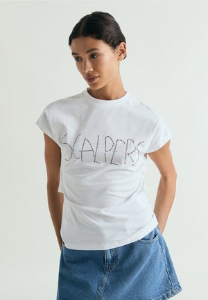 Mujer de cabello oscuro lleva una camiseta blanca con “SCALPERS” en letras de puntos y una falda vaquera azul, de pie contra un fondo liso.