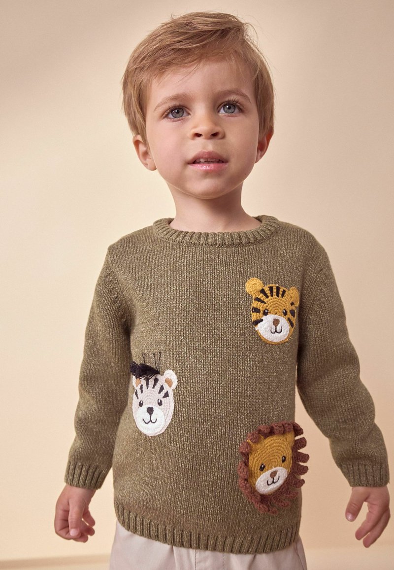 Lille barn iført en olivengrøn sweater med broderede dyreansigter, stående mod en ensfarvet beige baggrund.