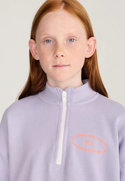 Jeune personne aux longs cheveux roux et aux taches de rousseur portant un haut zippé violet clair avec un texte orange sur la poitrine sur un fond uni.