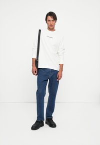 Hvit langermet skjorte med "Calvin Klein"-logo, blå jeans og svarte joggesko. Modellen bærer en svart skulderveske. Enkelt, uformelt utseende.