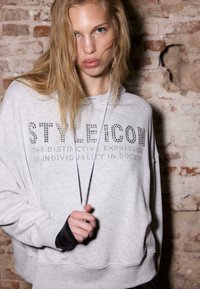 Szara bluza z kapturem, z troczkiem i napisem z kryształków "STYLE ICON". Ma luźny krój i paskowane mankiety na rękawach.