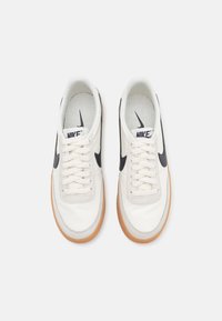 Valkoiset kangaskengät, joissa on musta Nike-logon kaarros, mantelinmuotoinen kärki, teksturoitu pohja ja beige kumipohja. Nauhat kulkevat kuuden reiän läpi.