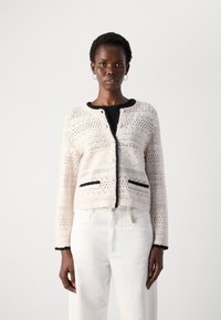 Cardigan lavorato a maglia color crema con intricati motivi a rete, bottoni neri e rifiniture. Presenta dettagli neri nelle tasche e un colletto rotondo.
