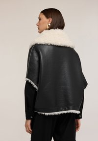 Kobieta w czarnej skórzanej kurtce z białym kołnierzem i wykończeniem z owczej wełny, zwrócona w lewo, na neutralnym tle.