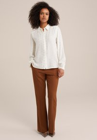 Camicia bianca con un motivo sottile, maniche lunghe e polsini elasticizzati, abbinata a pantaloni marroni su misura e scarpe a punta marroni chiare.