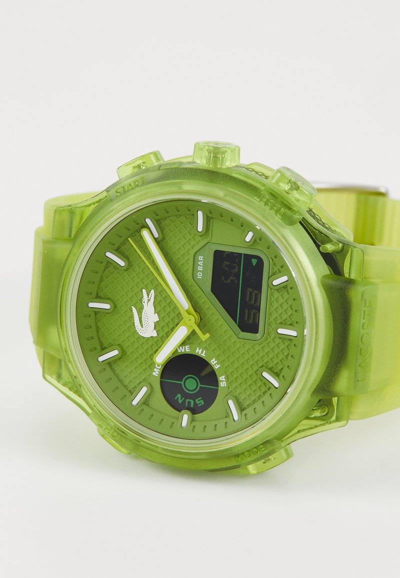 Reloj digital verde con esfera texturizada, caja de plástico transparente, acentos amarillos brillantes y correa de silicona. Presenta un logotipo y una pantalla digital.