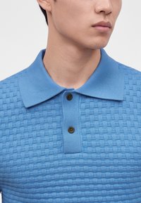 Homme portant un polo bleu clair texturé avec deux boutons fermés, visible du cou jusqu'au milieu de la poitrine.