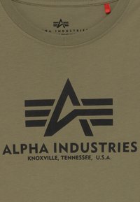 Olīvu zaļš T-krekls, izgatavots no kokvilnas, ar lielu melnu logo, kurā ir stilizēti burti un spārni, un zem tā ir teksts "ALPHA INDUSTRIES".