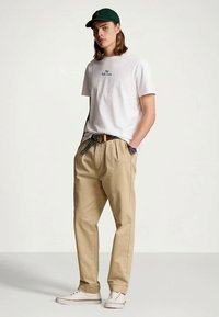 Νεαρός άνδρας με λευκό T-shirt Ralph Lauren, μπέζ πλισέ παντελόνι με ριγέ ζώνη, πράσινο καπέλο, λευκά αθλητικά παπούτσια και ρολόι, στέκεται με τα χέρια στις τσέπες.