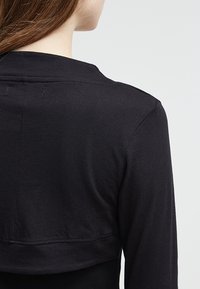 Svart kort jacka med en slät textur, långa ärmar och en enkel design med synliga sömnadsdetaljer vid halsringningen och fållen.