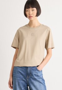 Donna con taglio di capelli bob corto, che indossa una camicia beige a maniche corte con un piccolo logo e jeans blu, con una mano in tasca, su uno sfondo neutro.