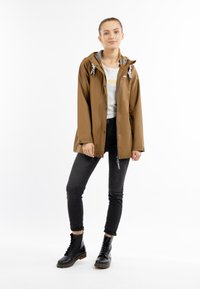 Giacca impermeabile marrone con cappuccio, foderata in grigio e con cordini, abbinata a jeans skinny neri e stivali neri stringati.