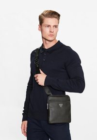 Sac bandoulière en cuir noir avec une texture lisse, forme rectangulaire, fermeture éclair et accent de logo. Porté en travers du corps.