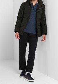 Zwarte pufferjack met een rits aan de voorkant, gedragen over een marineblauw henley-shirt. Gecombineerd met donkere broeken en marineblauwe sneakers met bruine accenten.