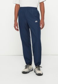 Moški nosi temno modre tekaške hlače Umbro in bež ter sivo športno obutev, stoji proti beli ozadju.