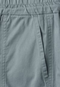 Gros plan d'un pantalon en tissu gris montrant une ceinture élastique et une poche latérale avec des détails de couture visibles.