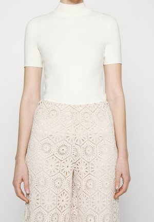 Kobieta ubrana w biały golf z krótkim rękawem wykonany z prążkowanego materiału oraz w beżowe, szydełkowane spodnie o szerokich nogawkach z wysokim stanem.