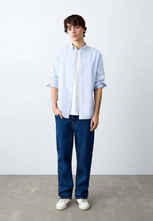 Hellblaues Hemd mit Knöpfen über einem weißen T-Shirt, kombiniert mit dunkelblauen Jeans und weißen Sneakers. Das Hemd hat hochgekrempelte Ärmel.