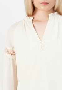 Donna che indossa una blusa crema con bordo in pizzo sul collo a V e inserti di pizzo trasparente sulle maniche lunghe, pelle chiara e capelli biondi lisci.