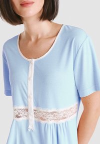 Top de loungewear azul claro con mangas cortas, que presenta un escote redondo, una parte frontal con botones y un detalle de encaje blanco en la cintura.