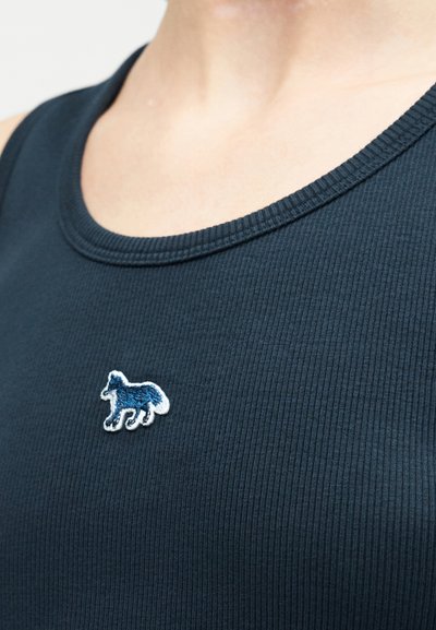 Άτομο που φοράει σκουρό μπλε ριμπ τοπ χωρίς μανίκια με ένα μικρό κεντητό μπλε και λευκό πατς σκύλου στο στήθος.