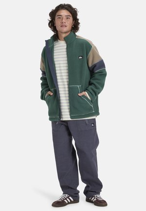 Groene fleece jacket met meerkleurige panelen, rits aan de voorkant, twee frontzakken, gelaagd over een gestreept shirt, gecombineerd met donkere broek en sneakers.