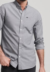 Graues Hemd mit Knopfleiste und kariertem Muster, einer einzelnen Brusttasche und hochgekrempelten Ärmeln. Hergestellt aus einem leichten, strukturierten Stoff.