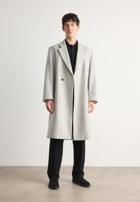 Šedý vlněný overcoat s dvouřadým designem, se zářezovým límcem, černými knoflíky, v kombinaci s černými kalhotami a tmavými botami.