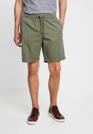 Mann trägt olivgrüne Shorts mit Kordelzug, graues T-Shirt und braune Ledersneaker mit weißen Sohlen, steht mit den Händen in den Taschen.