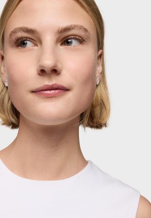 Młoda kobieta z krótkimi blond włosami, nosząca srebrne owalne kolczyki i biały bezrękawnik, patrząca lekko na bok.