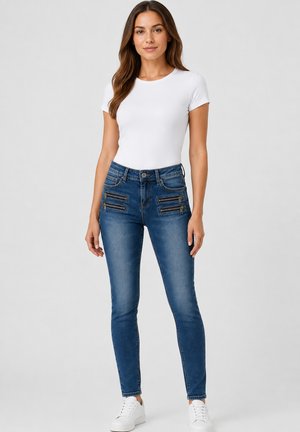 Kvinde står iført en hvid kortærmet top, blå jeans med fire forlommer med lynlås og hvide sneakers på en ensfarvet baggrund.
