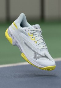 White athletic shoe with yellow accents, featuring a textured upper, prominent logo, and a lightweight design ideal for sports.