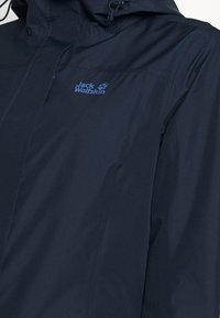 Veste imperméable bleu marine en tissu texturé, avec capuche ajustable et logo bleu "Jack Wolfskin" sur la poitrine. Fermeture avant zippée.