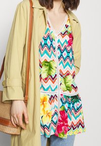 Top estivo colorato e a fantasia con motivi a zigzag e floreali. Caratterizzato da un bordo in pizzo e un design a strati, abbinato a un cappotto verde oliva chiaro e a una borsa di paglia.