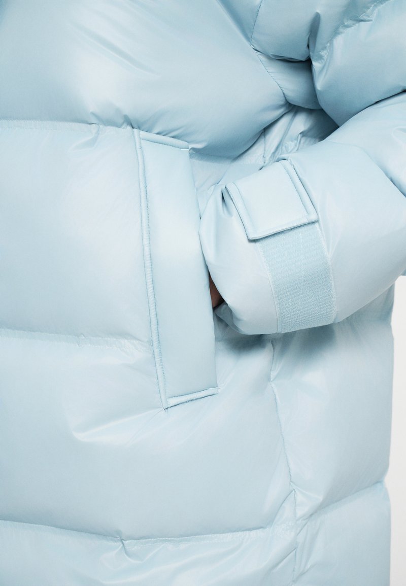 Détail d'une veste doudoune bleu clair avec une poche latérale et un poignet en velcro, présentant une texture lisse et brillante et un rembourrage duveteux.